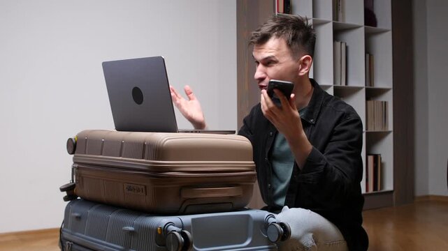 Man sitting by stacked suitcases, actively using a laptop for planning and speaking into a smartphone, combining work and personal communication before a trip