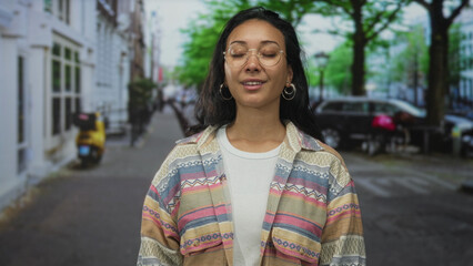 Woman with eyes closed, wearing glasses, hoop earrings and a patterned jacket on a tree lined street with parked cars  calm contentment reflection. © Krakenimages.com