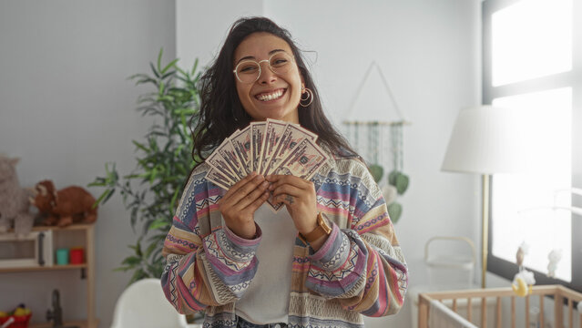 Woman holding a fan of cash with hands visible in a baby room inside a residential building; financial joy.