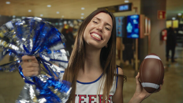 Young hispanic cheerleader holds blue pompom and football while smiling in airport terminal; excitement.