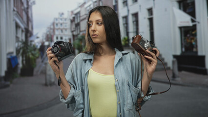 Fototapeta premium Woman holds two cameras in each hand on a city street while looking at the viewer, denim shirt visible; confidence exploration.
