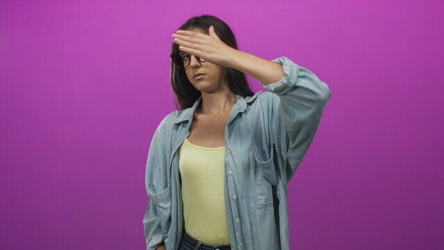 Young hispanic woman covering eyes with hand in studio against purple wall wearing denim shirt and yellow tank; shyness solitude.