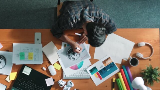 Top view of designer writing and designing logo graphic at meeting. Closeup of business man working on colored palettes and writing idea on sticky notes on table with equipment and paper. Symposium.