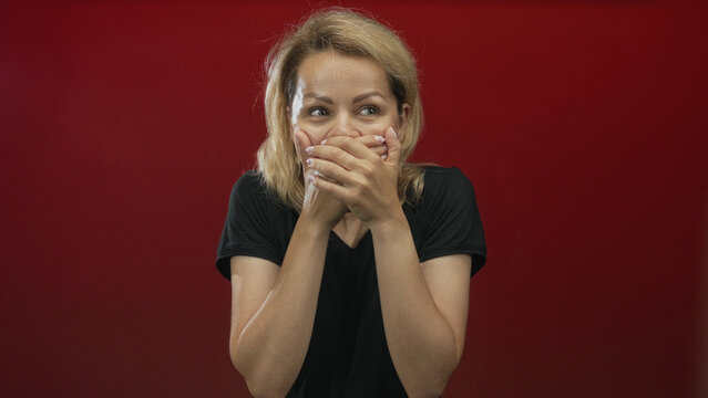 Blonde woman in black shirt with wide eyes, hands covering mouth and staring at camera in studio; surprise.