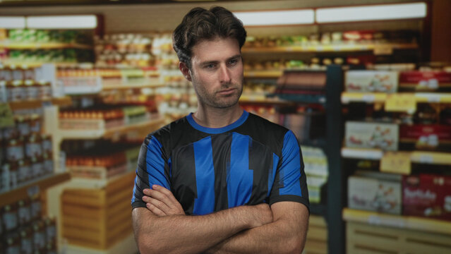 Man with arms crossed showing forearms in supermarket aisle by stocked shelves and packaged groceries, looking to the side; confidence.
