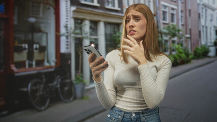 Fototapeta premium Woman holding smartphone with hand on chin gesture on a narrow city street with bicycles and storefronts visible; thoughtful hesitation.