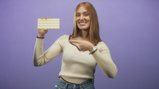 Young woman holding white chocolate bar points finger toward camera in studio; playful product promotion campaign.