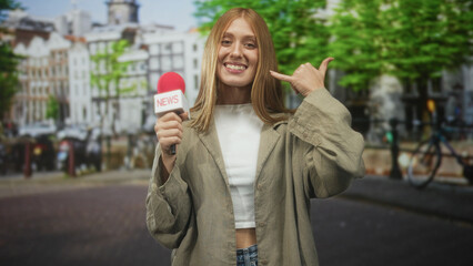 Fototapeta premium Woman redhead reporter holds news microphone and makes call me hand gesture on street by canal buildings; cheerful reporting.