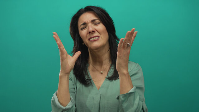 Middle aged woman raises hands to face in teal studio setting with expressive gesture and closed eyes showing despair; frustration.