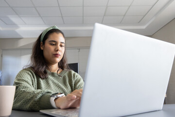 Woman wearing green knit sweater, headband watch typing on silver laptop at office table, beige mug