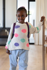 African American child standing at home holding banister wearing cream dot sweater purple backpack