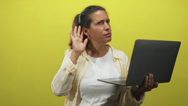 Middle age brunette woman holding a laptop with one hand and cupping her ear with the other while squinting at the screen in studio with yellow backdrop; curiosity.