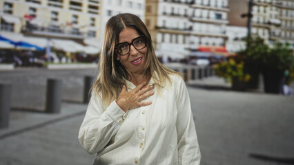 Fototapeta premium Woman wearing glasses with hand on chest in a busy street with buildings and cafe planters; gratitude reflection.