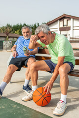 Fototapeta premium vertical Two active senior men on a bench smiling and wiping sweat while resting with a basketball and water bottle, enjoying sport and demonstrating healthy aging