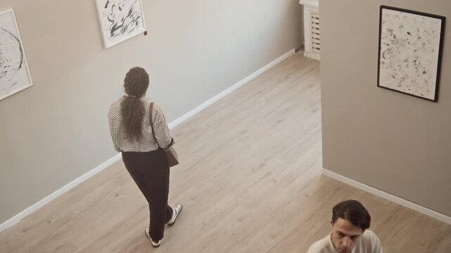 High angle shot of Caucasian gallery visitors of different age and gender walking in exhibition hall and observing framed abstract art paintings