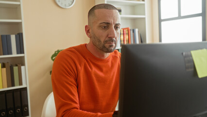 Fototapeta premium Man typing on computer at desk in office building, orange sweater and smartwatch visible, leaning toward monitor and focused on screen; concentration focus.