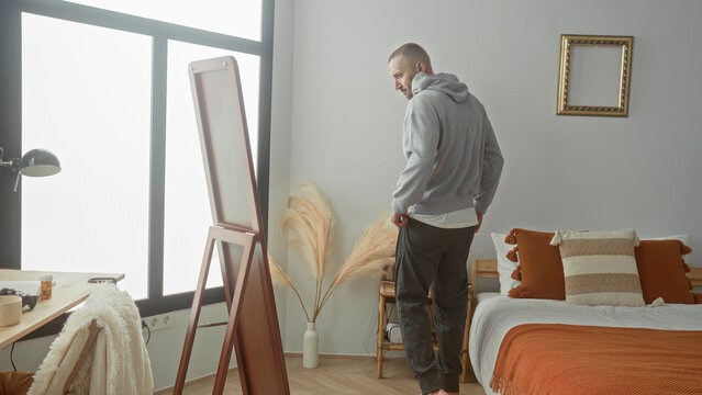 Man pulls hoodie over head by bed and mirror, adjusts hood and checks face in full length mirror in a sunlit bedroom building; calm reflection.