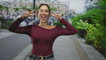 Fototapeta premium Woman shows peace sign with both hands, bare shoulder visible and smiling on city street with plants and buildings; joyful.