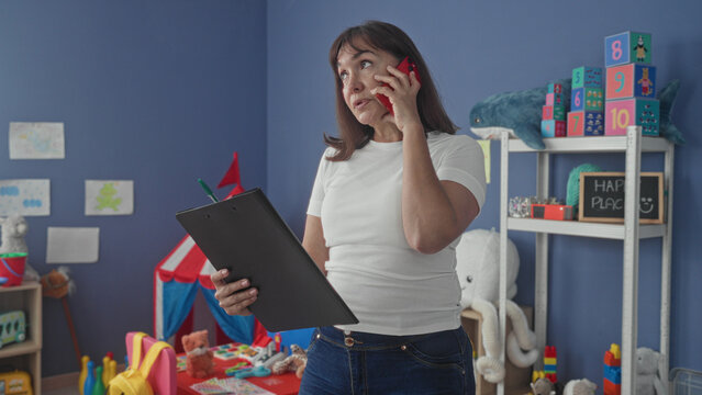 Woman middleaged teacher holds clipboard and phone to ear, checking notes with red smartphone amid toys on shelves in building classroom; concern responsibility.