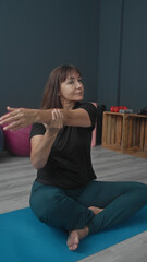 Middle aged woman seated crosslegged holds forearm across chest on blue yoga mat in studio;...