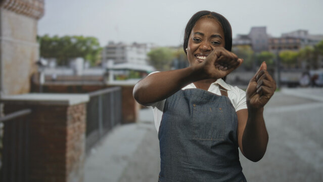 Woman in denim apron smiling and miming a pinching motion with her hands on a street; joy hospitality.