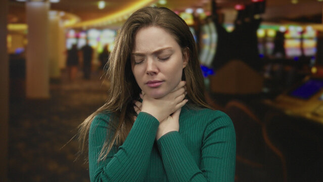 Woman clutching her throat while standing before a row of slot machines in a casino building; panic.