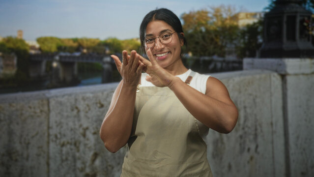 Woman in apron claps hands on street by stone bridge parapet while smiling and adjusting glasses; joy warmth friendliness.