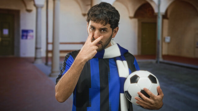 Man holding soccer ball points two fingers to face in building corridor wearing blue black team jersey and scarf; determination fan support.