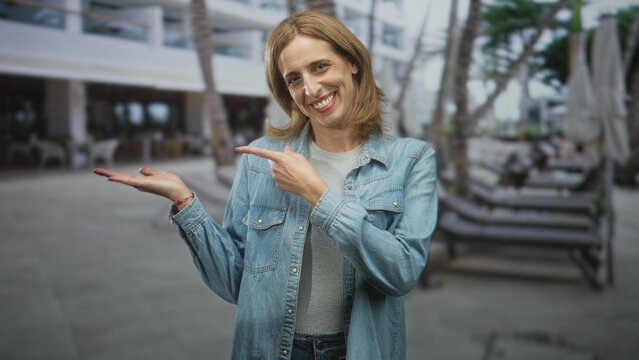 Woman in denim jacket points with open palm to poolside lounge chairs beside a hotel building, smiling and slightly turned to the side; inviting welcome.