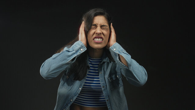 Woman covering ears with hands in studio against black backdrop wearing striped top and denim jacket; sensory overload distress.