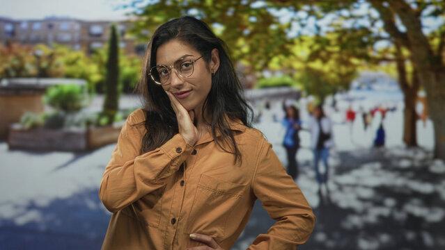 Woman wearing glasses with hand on chin and elbow akimbo in urban street plaza with trees and blurred pedestrians; thoughtful reflection.