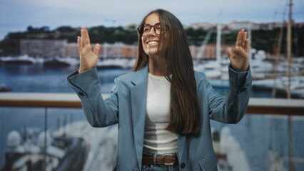 Young woman raising both hands with palms visible and smiling on a building balcony, wearing...