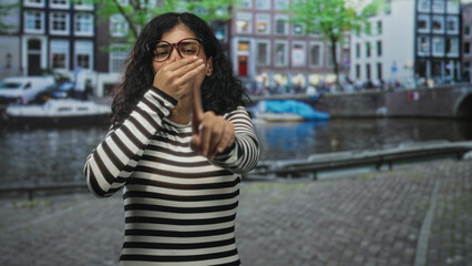 Fototapeta premium Young brunette woman covering mouth with hand and pointing finger on cobblestone street by amsterdam canal; silence warning.