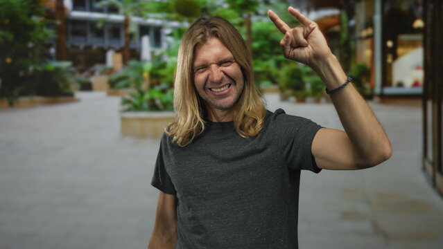 Man with long blond hair grinning widely raising both arms to display rock horns gesture on street; rock fun metal.