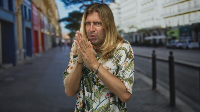 Blond man with long hair covers nose with hand on city street sidewalk near bollards amid empty storefronts; disgust.
