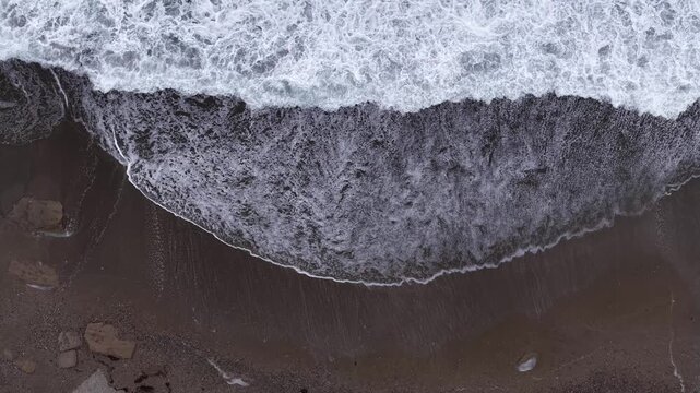 A serene aerial shot of the foamy waves meeting the dark-colored sand along the coast of Gazipaşa, Antalya. An aesthetically pleasing drone video for nature and ocean-themed projects.