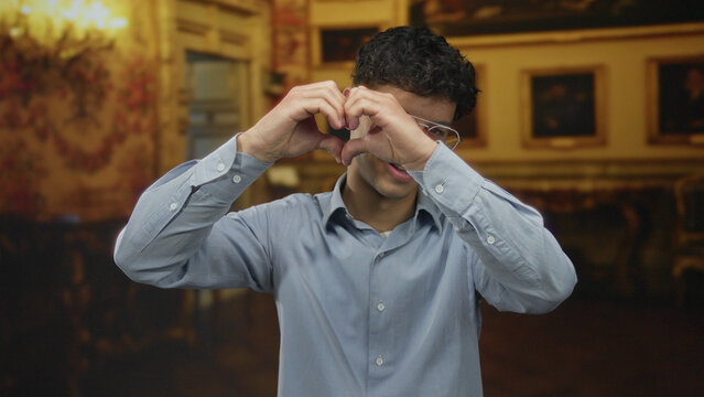 Young hispanic man wearing glasses making heart shape with hands inside ornate museum building hallway; affection.