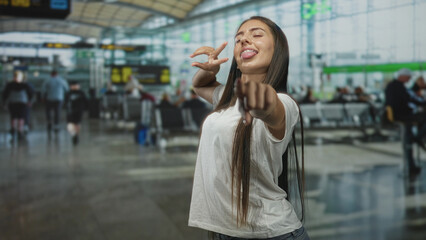 Fototapeta premium Young woman points finger at camera, tongue out showing braces, wearing white t shirt in an airport terminal; playful travel moment.