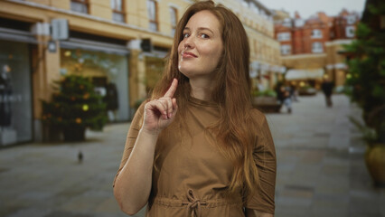 Fototapeta premium Young woman pointing finger to building on street under overcast sky among pedestrians next to storefront windows; attention.