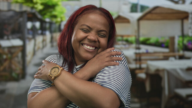 Woman smiling warmly hugging herself on a sunny outdoor restaurant terrace with wooden tables visible; joy self love comfort.
