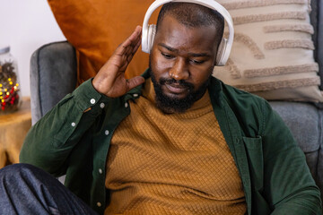 African American man sitting on gray couch at home, touching headphones, wearing mustard sweater