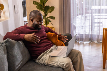 African American adult sitting on sofa in living room wearing maroon knit gesturing toward laptop
