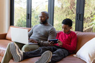 African American father and child sitting on sofa in living room using laptop and handheld device