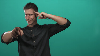 Fototapeta premium Man pointing finger at camera while touching temple with index finger in turquoise studio, wearing black shirt and smirking; confidence.