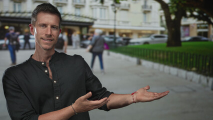Young caucasian man winks and presents open palms at a building entrance on a street; friendly...