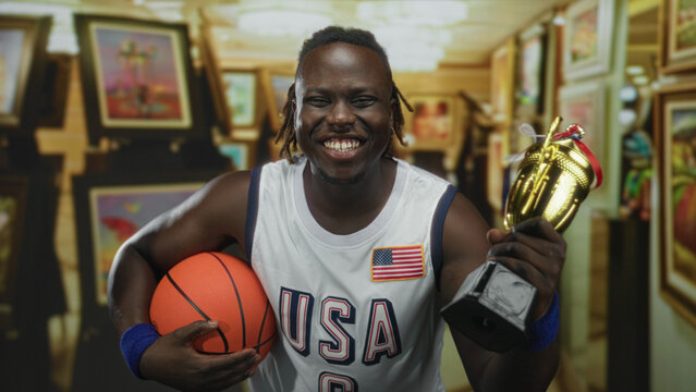 Man in usa basketball jersey holding basketball and trophy, smiling with teeth visible in gallery building surrounded by framed paintings; pride victory.