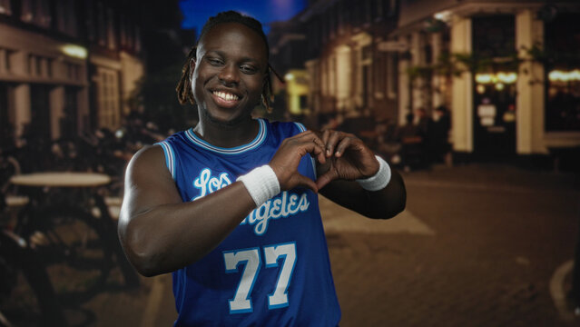 Man forms heart with hands on a bustling city street at night in blue jersey, smiling and posing by cafe tables; love connection.