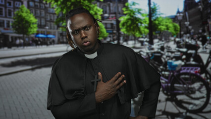 Fototapeta premium Man priest with hand on chest holding wooden cross, wearing clerical collar on busy city street with bicycles and trees; devotion prayer.