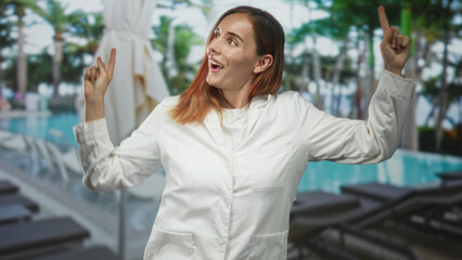 Fototapeta premium Woman doctor in white coat points index fingers up at a resort pool with lounge chairs and palm trees, smiling and eyes closed; playful confidence.