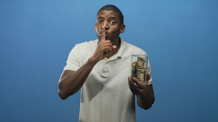 Young man holds glass jar of bundled dollar bills and presses finger to lips for silence in studio  secrecy. © Krakenimages.com